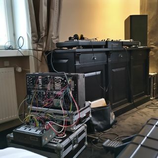 A fancy hotel room with two patched Eurorack cases sitting on a hardened audio gear transport crate. Behind, a DJ setup with two turntables, a mixer and a monitor speaker.